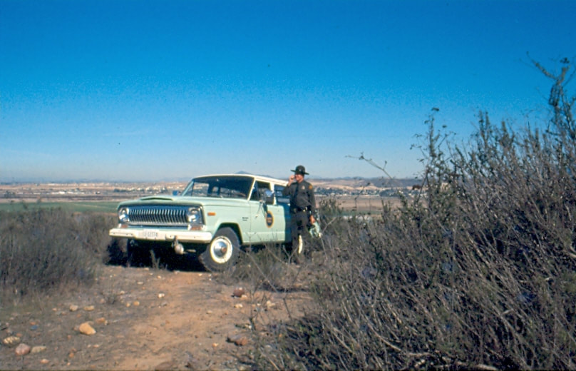 USBP Border Patrol photographs 1970-1990 an agent in dress uniform in the field next to a sea foam green SUV