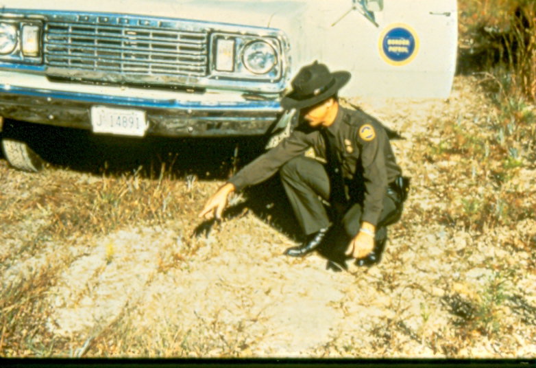 USBP Border Patrol photographs 1970-1990 an agent in dress uniform checking for sign in front of a sea foam green SUV